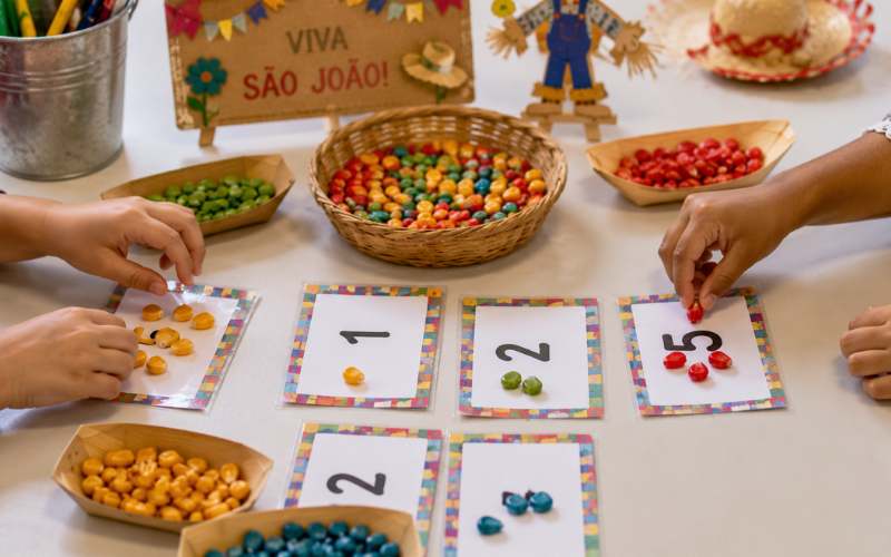 Mãos de crianças pequenas organizando grãos de milho coloridos em pratos, ao lado de cartões com números, em atividade de contagem com tema junino